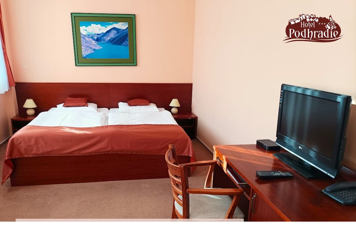 A hotel room with a bed, a desk with a TV, a chair, a nightstand with a lamp, and a framed picture on the wall.