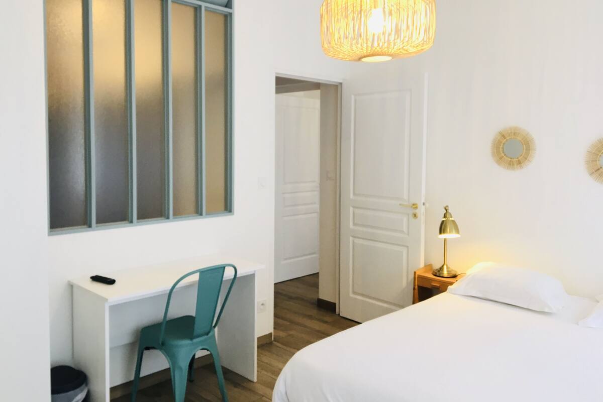 A hotel room with a bed, a desk with a chair, a hanging lamp, and a door leading to another room.