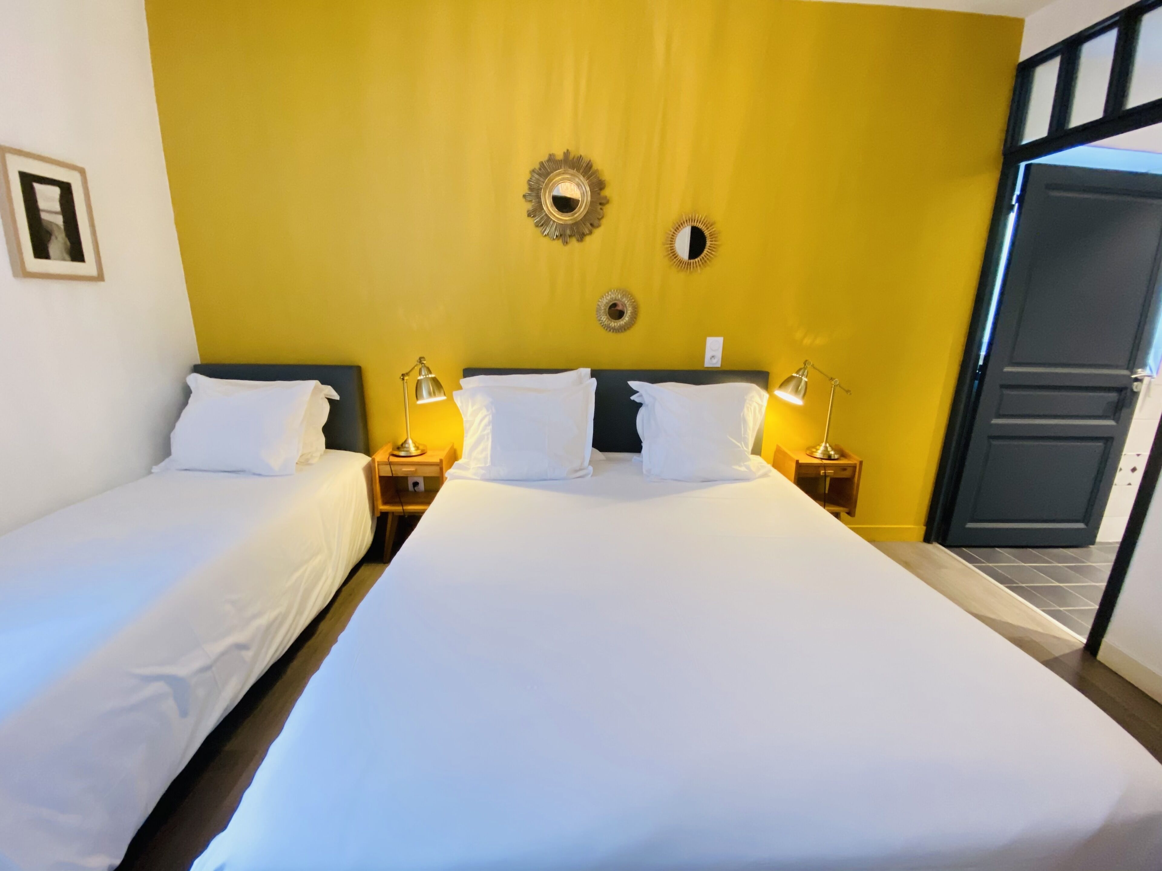 Une chambre d’hôtel avec deux lits, chacun agrémenté de draps blancs et d’oreillers assortis. On y trouve des tables de chevet avec des lampes, un miroir et un luminaire fixé au mur.