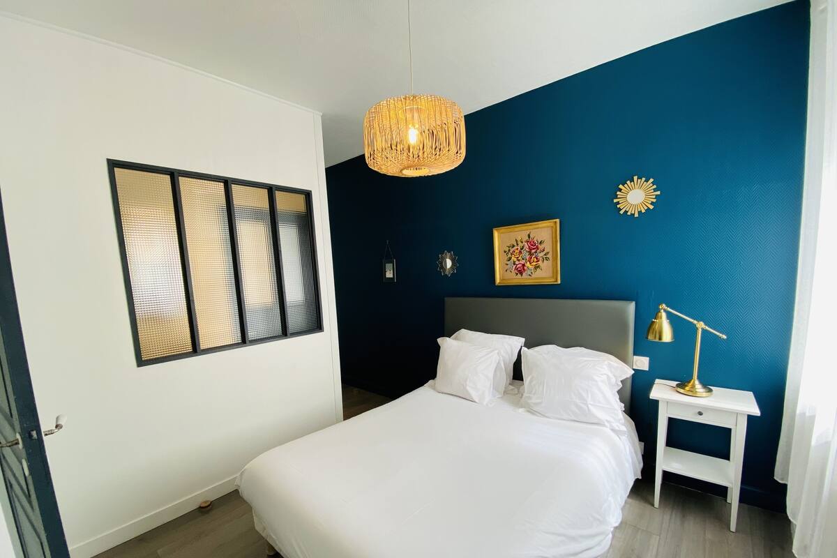 A neatly arranged bedroom with a large bed, a bedside table with a lamp, a hanging light fixture, a window with blinds, and a wall-mounted picture.