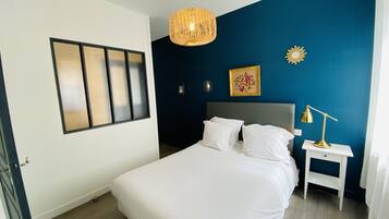 Un dormitorio ordenado con una cama grande, una mesita de noche con lámpara, una lámpara colgante, una ventana con persianas y un cuadro en la pared.