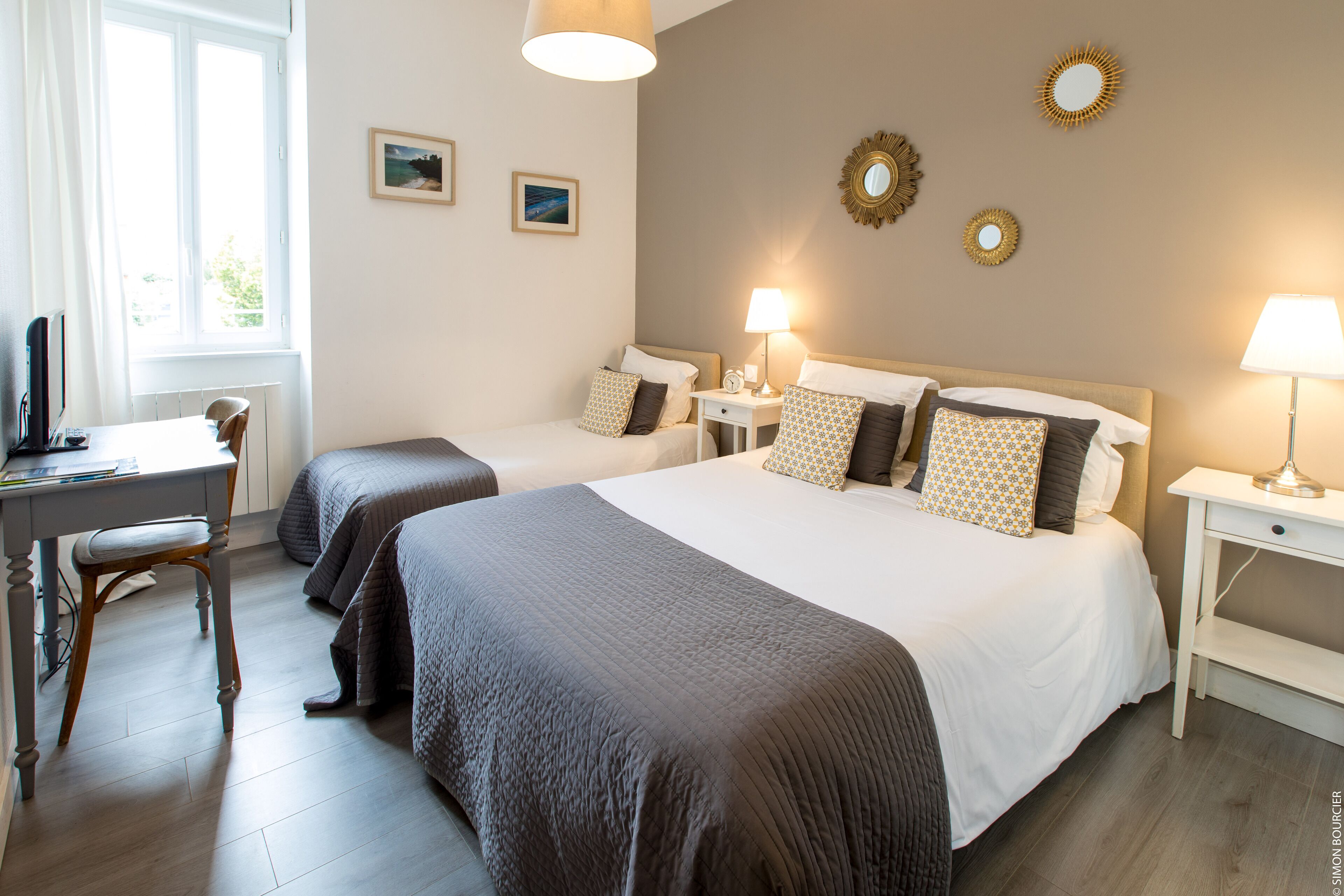 A hotel room with two beds, a desk, and a chair. There are wall-mounted lamps and picture frames.