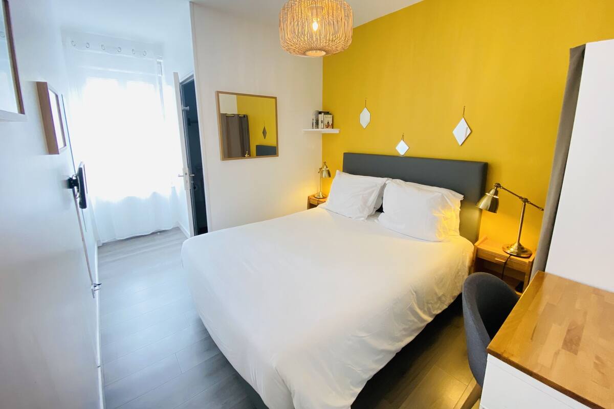 A modern hotel room with a large bed, a desk, and a chair. There is a mirror on the wall and a hanging light fixture.