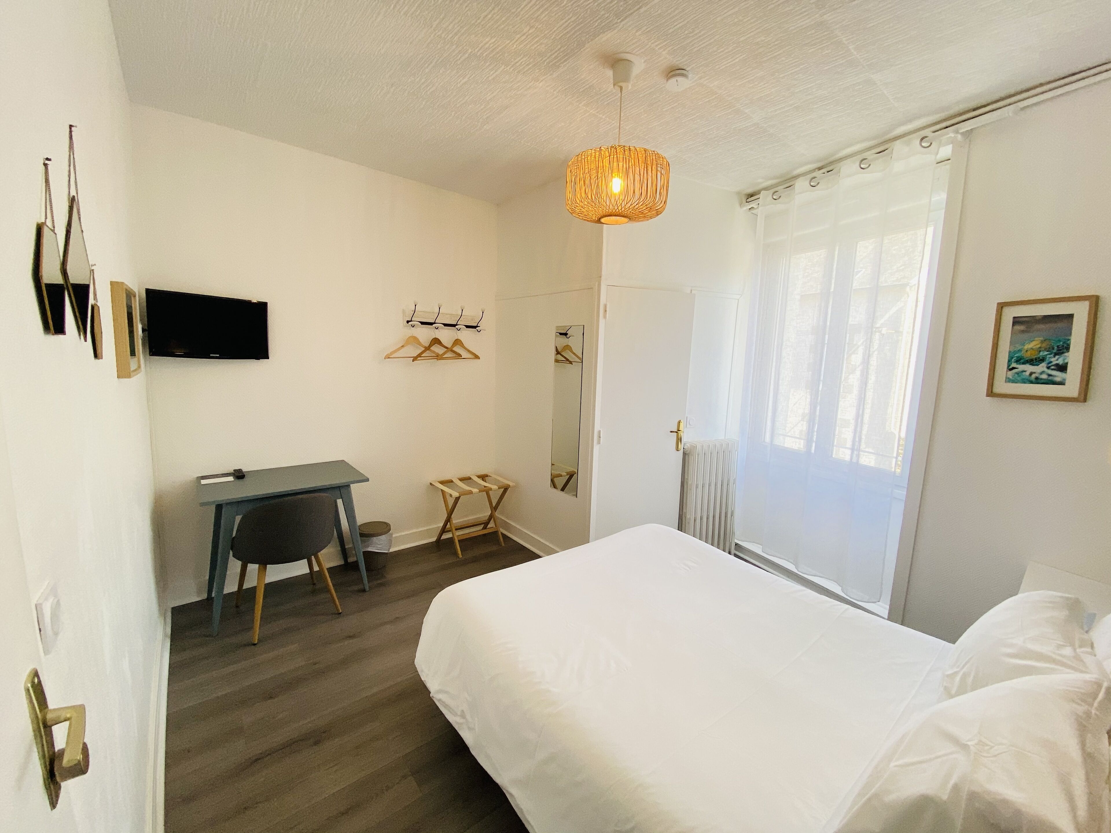 Une chambre à coucher comprenant un lit, un bureau, une chaise, un luminaire suspendu et une fenêtre avec des rideaux.