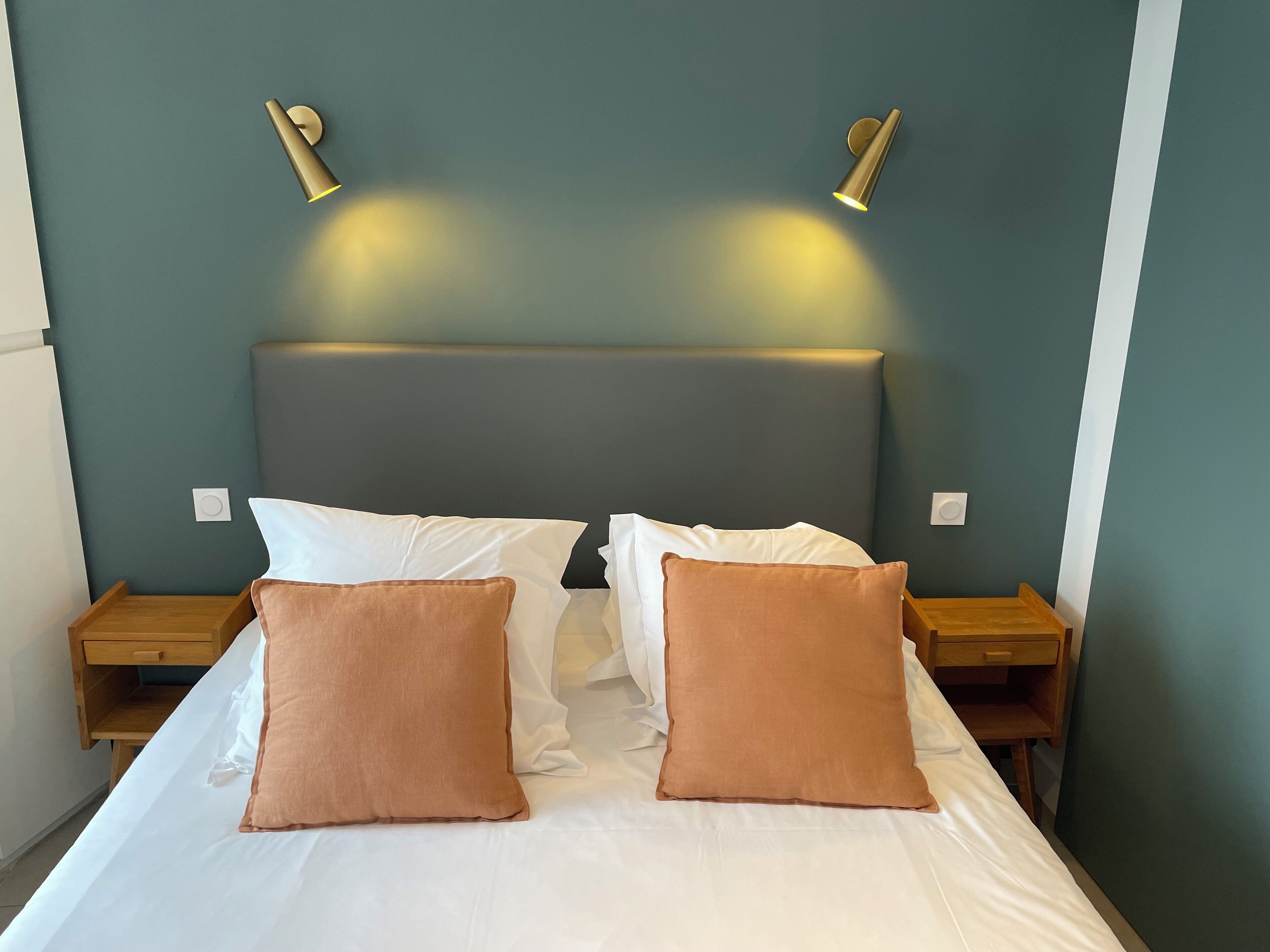 Cama bem arrumada com dois travesseiros brancos e dois marrons, acompanhada por mesas de cabeceira de madeira com gavetas, em um quarto com paredes verde-água e cabeceira cinza.