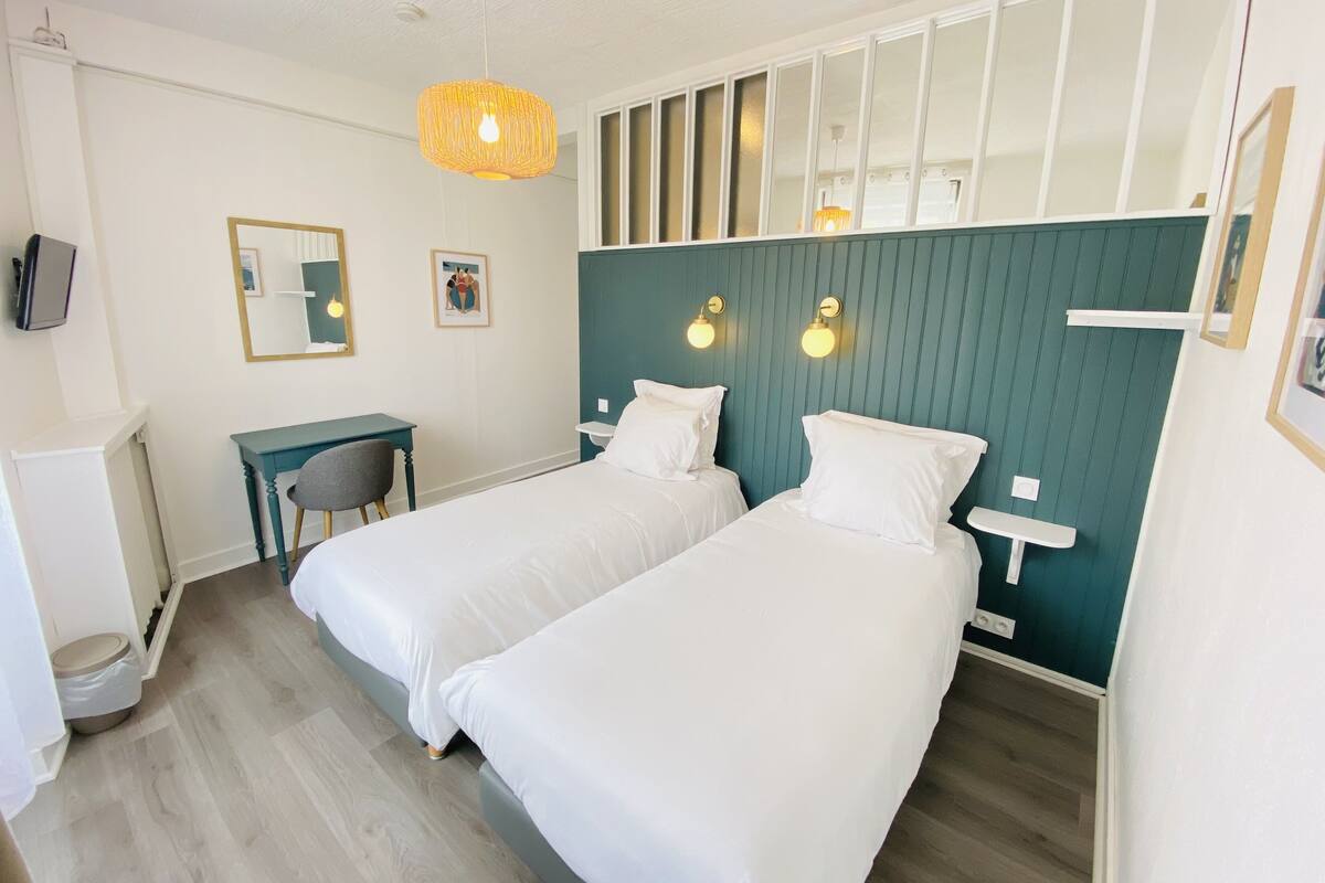 A hotel room with two beds, a desk, and a chair. There are wall-mounted lights and a hanging light fixture.