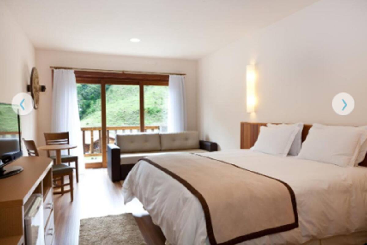 Een slaapkamer met een bed, een bureau en een stoel. Er is een raam met gordijnen en uitzicht op groen.