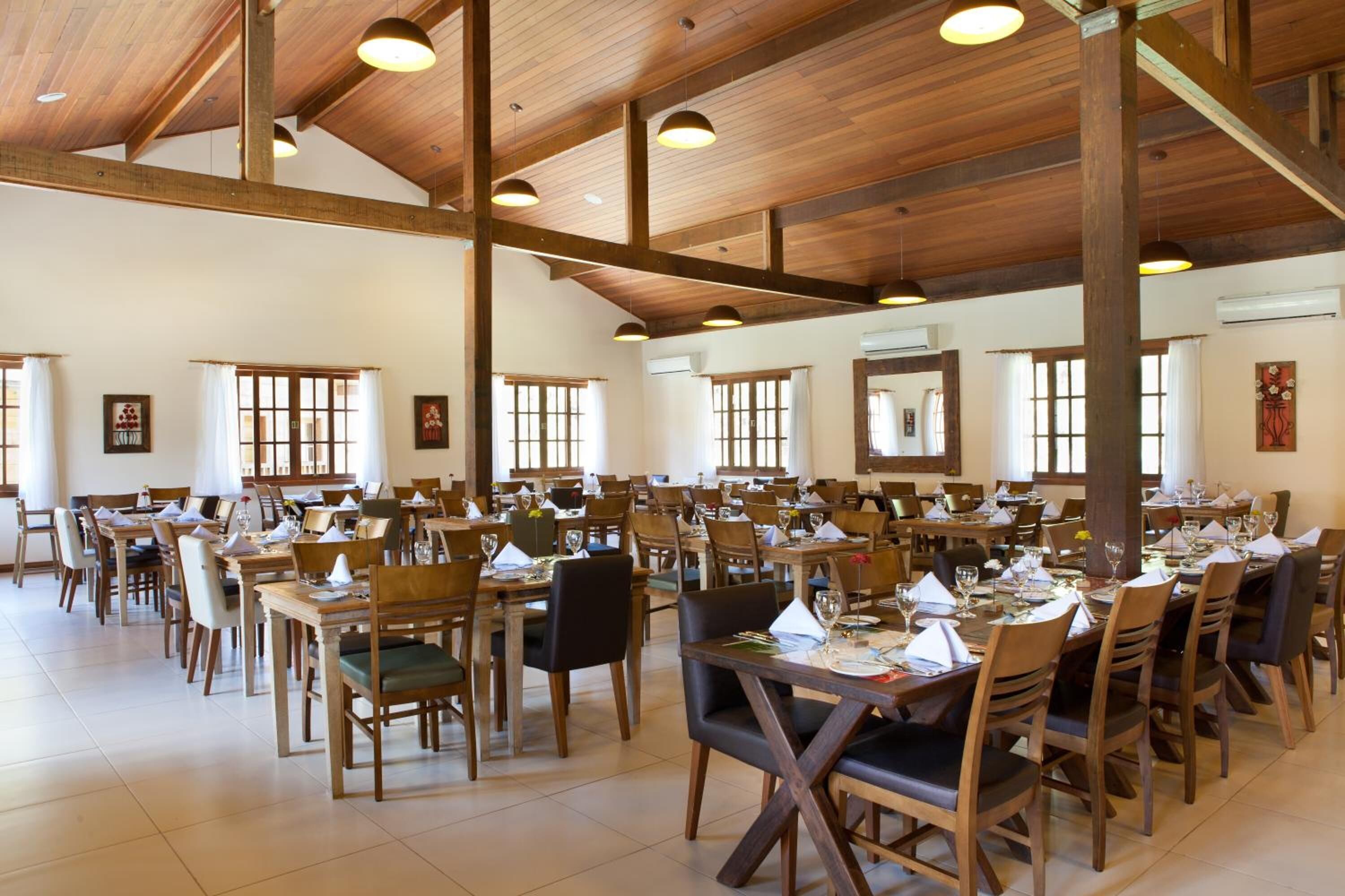 A spacious dining hall with wooden tables and chairs, large windows, and a high wooden ceiling.