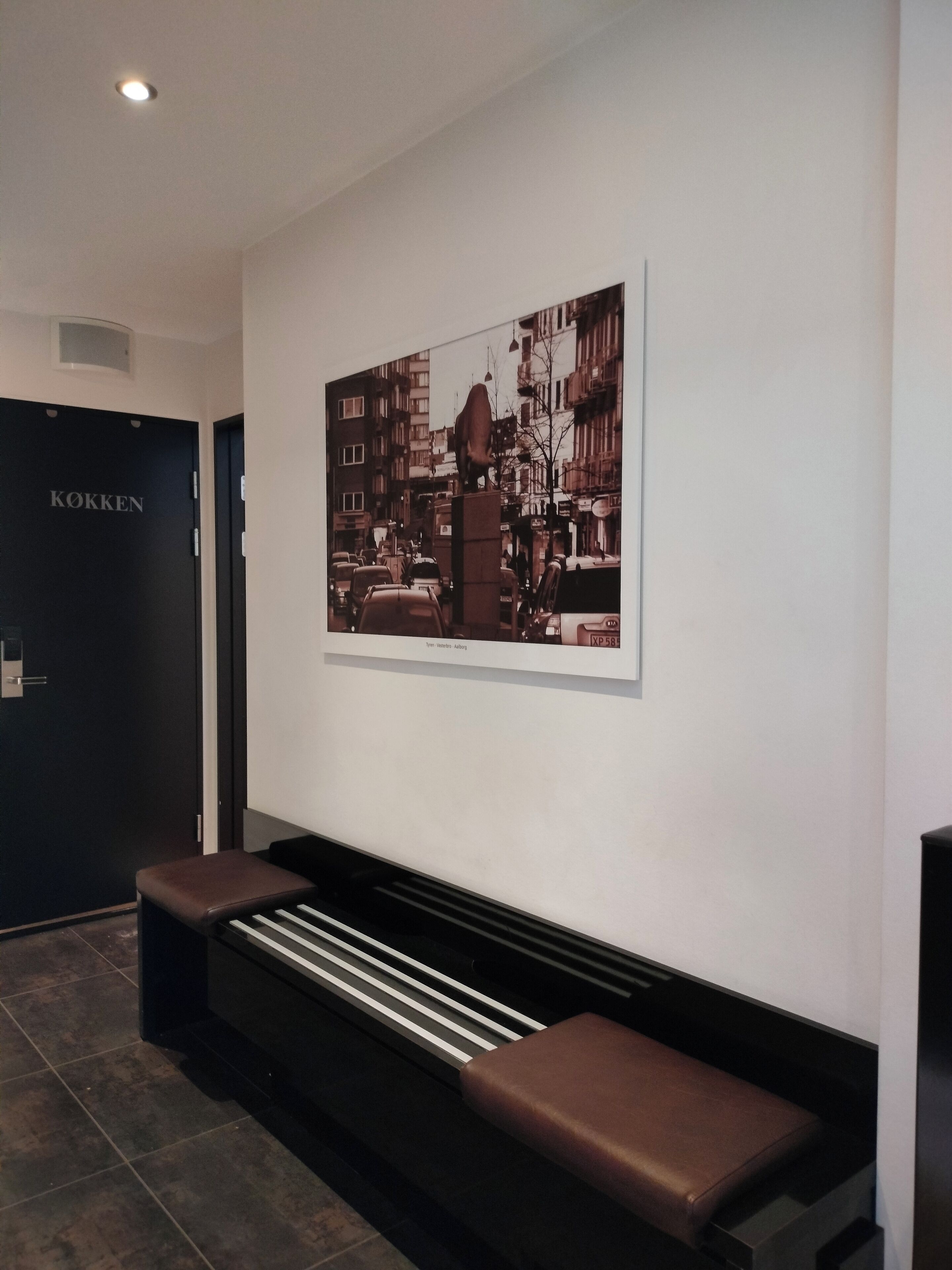 Un interior moderno con un banco negro, una obra de arte urbana enmarcada y una puerta con la leyenda "KOKKEN".