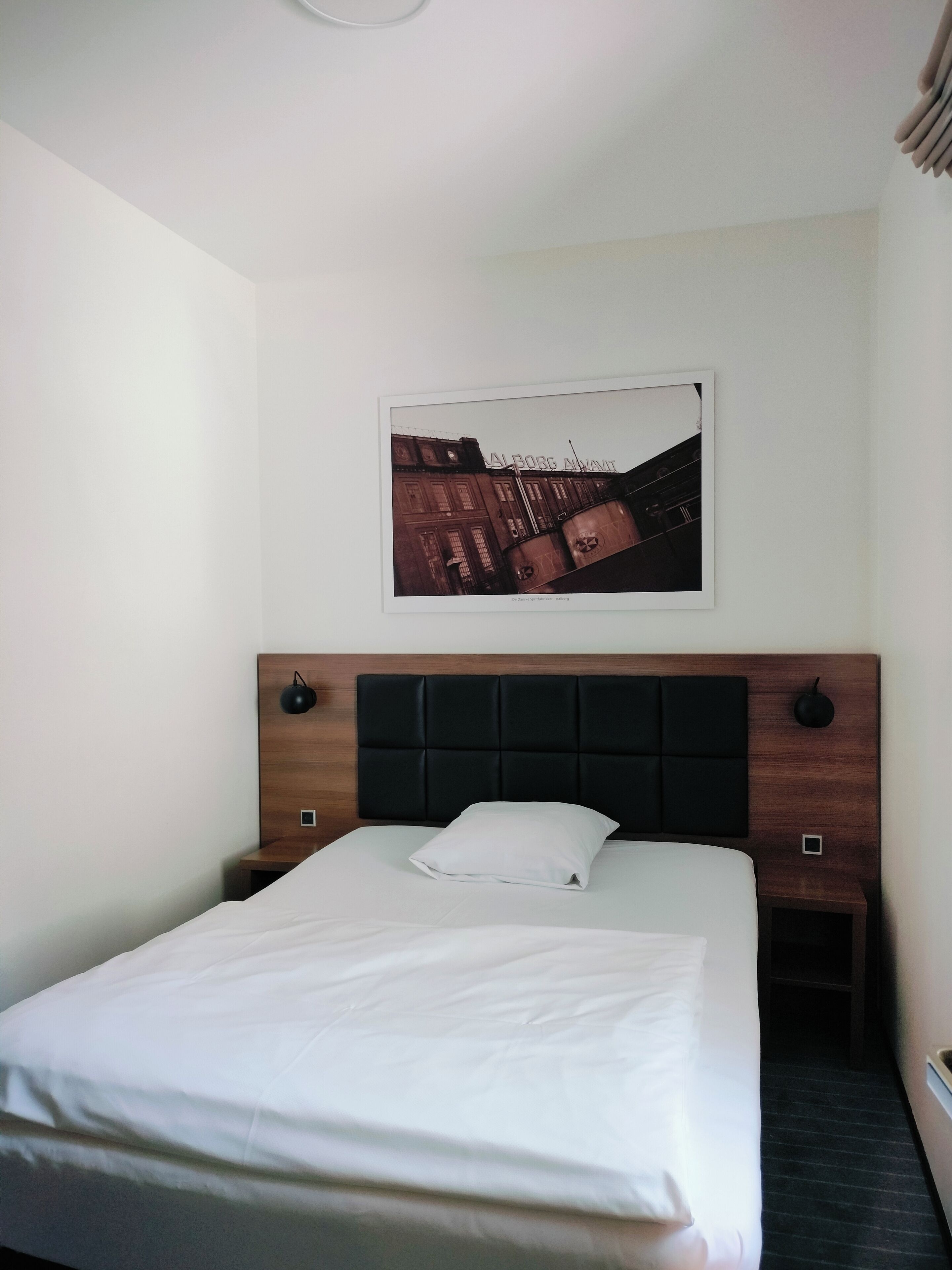 A hotel room with a neatly made bed, a headboard with a dark panel, a framed picture on the wall, and a small bedside table with a lamp.