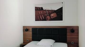 A hotel room with a neatly made bed, a headboard with a dark panel, a framed picture on the wall, and a small bedside table with a lamp.
