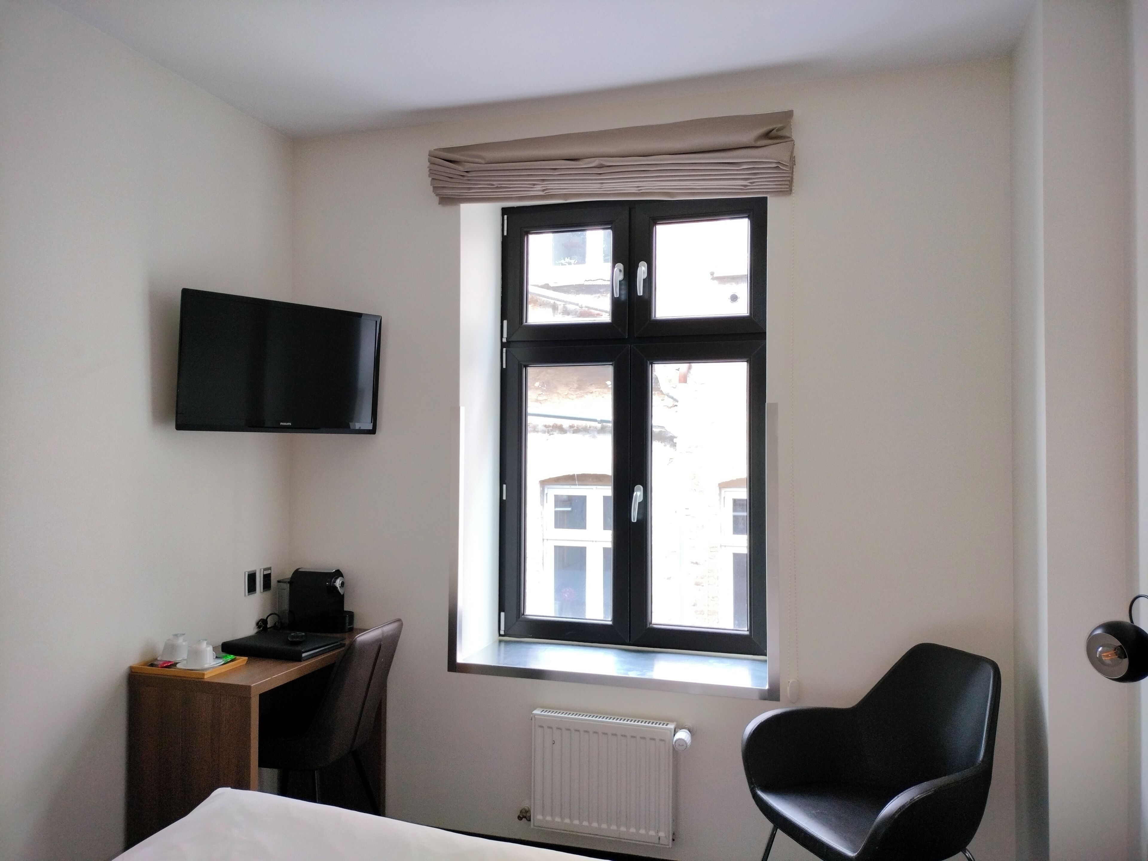 A hotel room with a bed, a desk with a chair, a television mounted on the wall, and a window with a view of buildings outside.