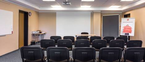 Un salón de conferencias con filas de sillas, un proyector, una pizarra blanca y un cartel.