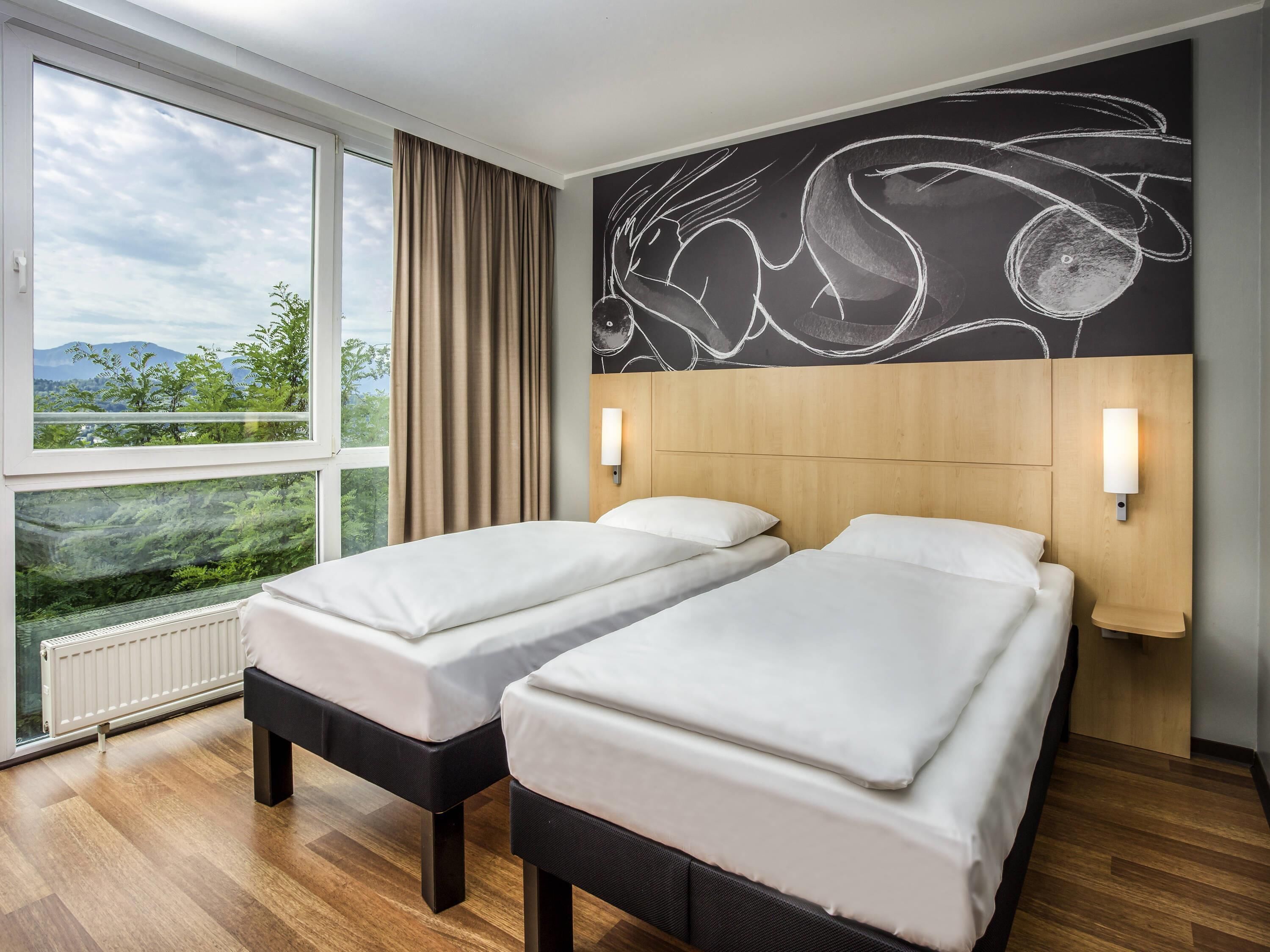 A hotel room with two beds, a large window with a view of greenery and mountains, a wall with abstract art, and a wooden headboard.