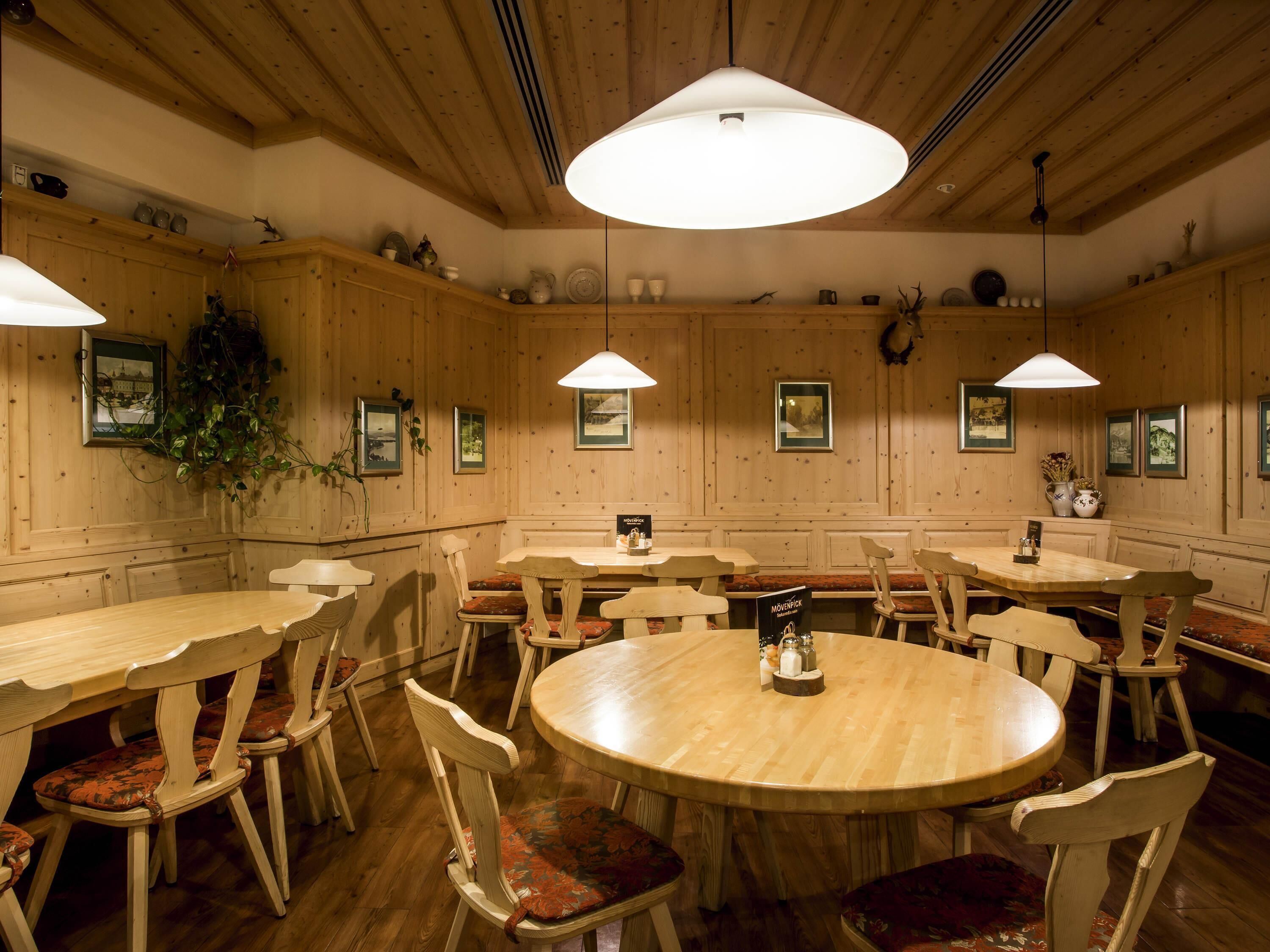 A wooden interior with round tables and chairs, pendant lighting, framed pictures on the walls, and a hanging plant.