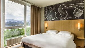 Habitación de hotel con una cama grande, dos lámparas de noche, vistas a montañas y vegetación a través de una ventana, y una obra de arte moderna y abstracta en la pared.