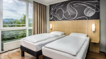 Une chambre d’hôtel avec deux lits, une grande fenêtre donnant sur un paysage verdoyant et des montagnes, un mur orné d’œuvres d’art abstrait et une tête de lit en bois.