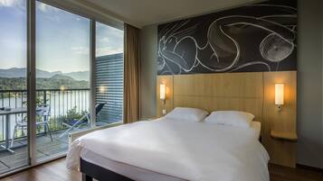 Habitación de hotel con una cama grande, dos lámparas de noche, vista a un lago y un balcón con mesa y sillas.