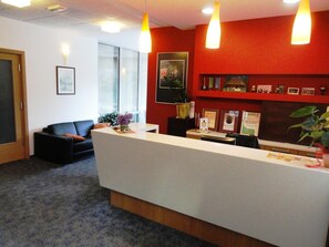 A reception area with a white reception desk, a black sofa, and a red accent wall. - Hotel Bela Krajina (Metlika)