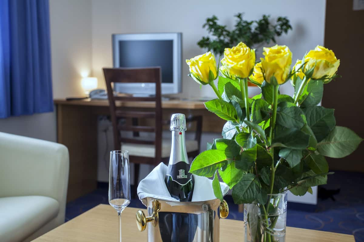 Une chambre avec un lit, un bureau avec un ordinateur, une chaise, un vase contenant des roses jaunes et une bouteille de champagne.