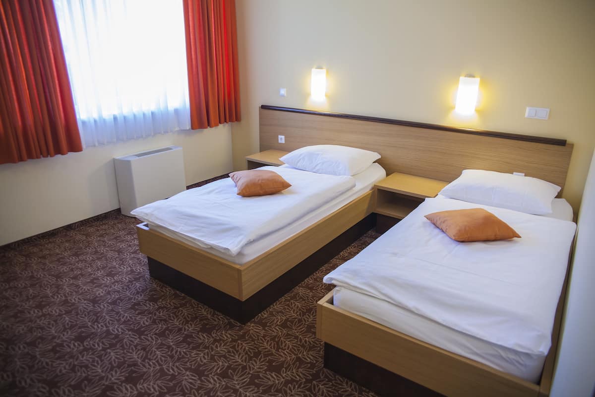Une chambre d’hôtel avec deux lits, chacun recouvert de draps blancs et agrémenté d’un seul oreiller orange. On y trouve des tables de chevet avec des lampes, un radiateur et une fenêtre avec des rideaux rouges.