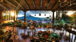 An outdoor dining area with a view of the ocean and mountains, featuring rustic wooden beams, metal chairs, and a decorative wreath. - Hotel Atitlán (Panajachel)