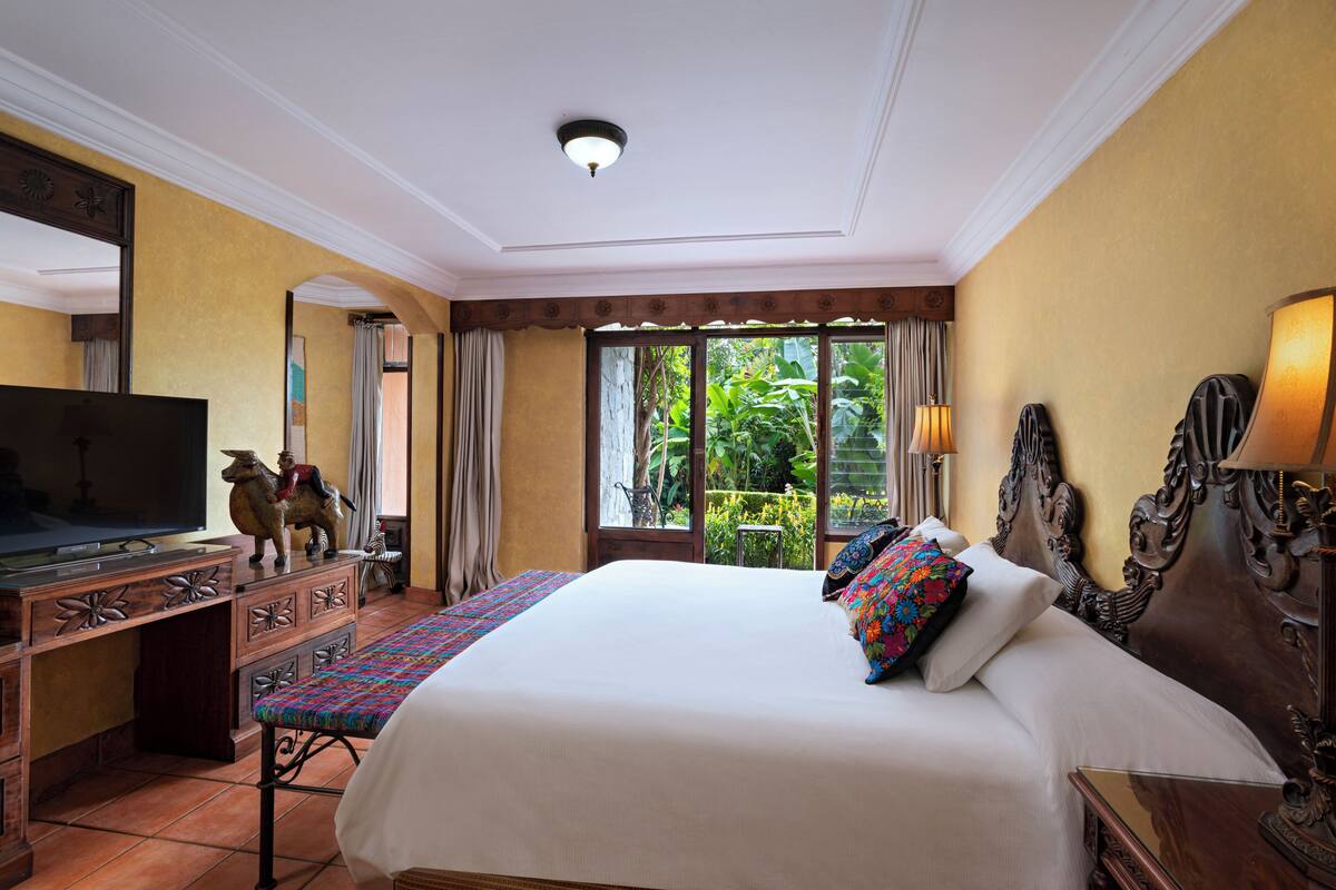 Habitación de hotel con una cama grande, televisor, escritorio con cajones y vista a la vegetación por la ventana.