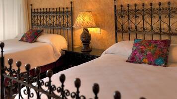 Habitación de hotel con dos camas, cada una con almohada floral, una mesita de noche con lámpara y cabeceros de hierro forjado.