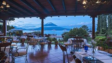 An outdoor dining area with tables and chairs, overlooking a body of water and a mountain.