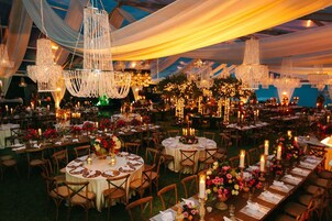 Un salón interior para eventos con mesas redondas dispuestas para comer, candelabros colgando del techo y centros de mesa florales.