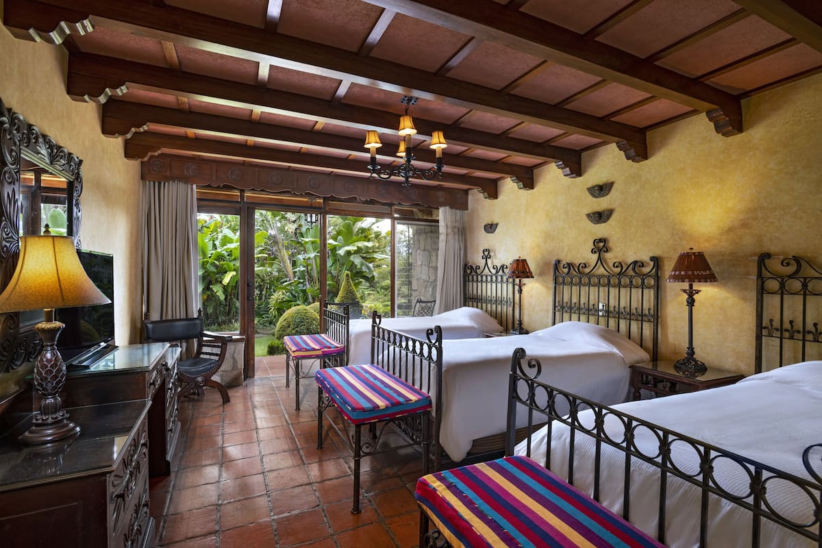 Habitación de hotel con dos camas, un escritorio con lámpara y vistas a zonas verdes.