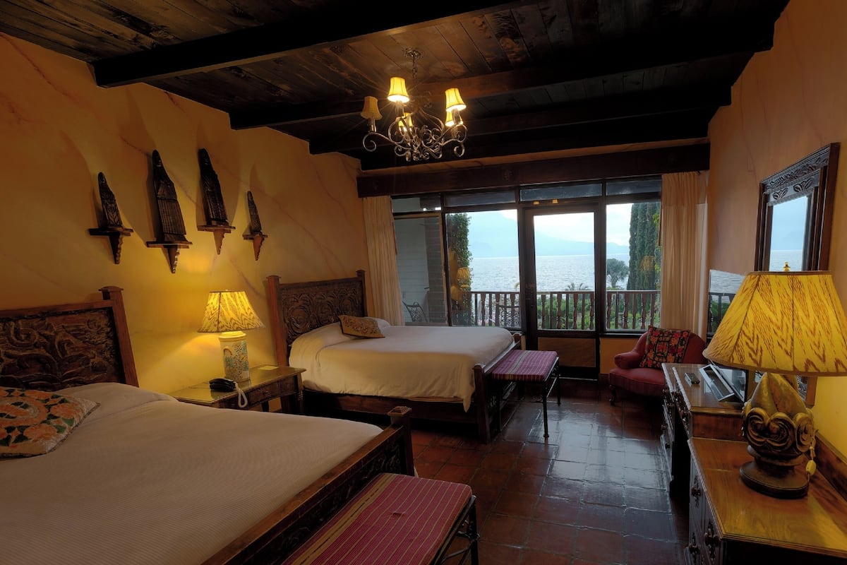 Habitación de hotel con dos camas, techo de madera, araña, vista al mar y balcón con mesa y silla.