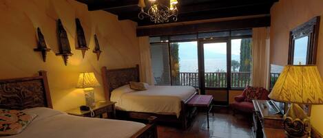 A hotel room with two beds, a wooden ceiling, a chandelier, a view of the sea, and a balcony with a table and chair.