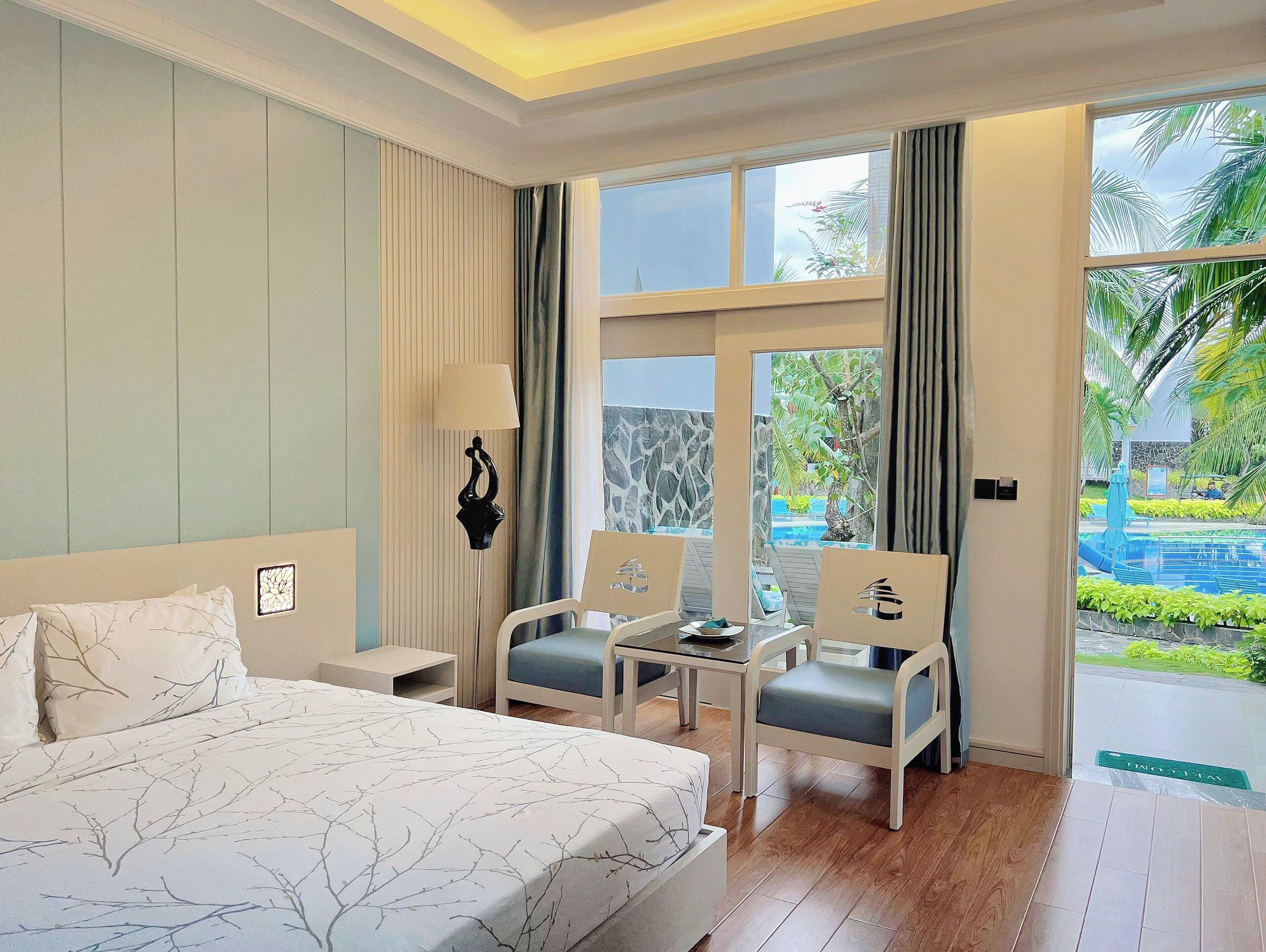 Habitación de hotel con cama, dos sillas, una mesa y un ventanal con vistas a una piscina y follaje.
