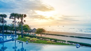 A poolside view with a beach and ocean in the background, palm trees, and thatched umbrellas.