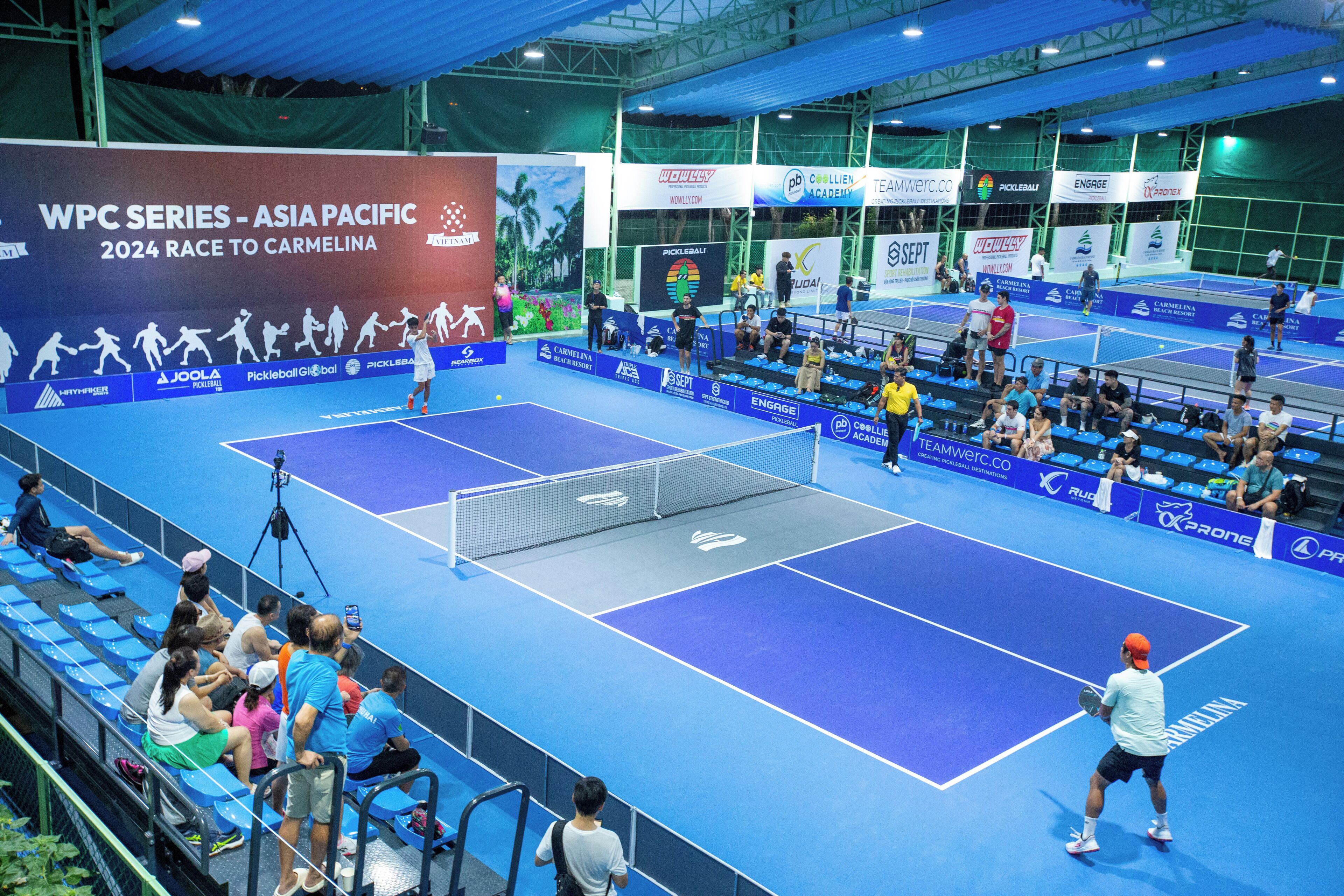 A tennis court with blue flooring and white netting, surrounded by blue seating areas and a large banner with event details.