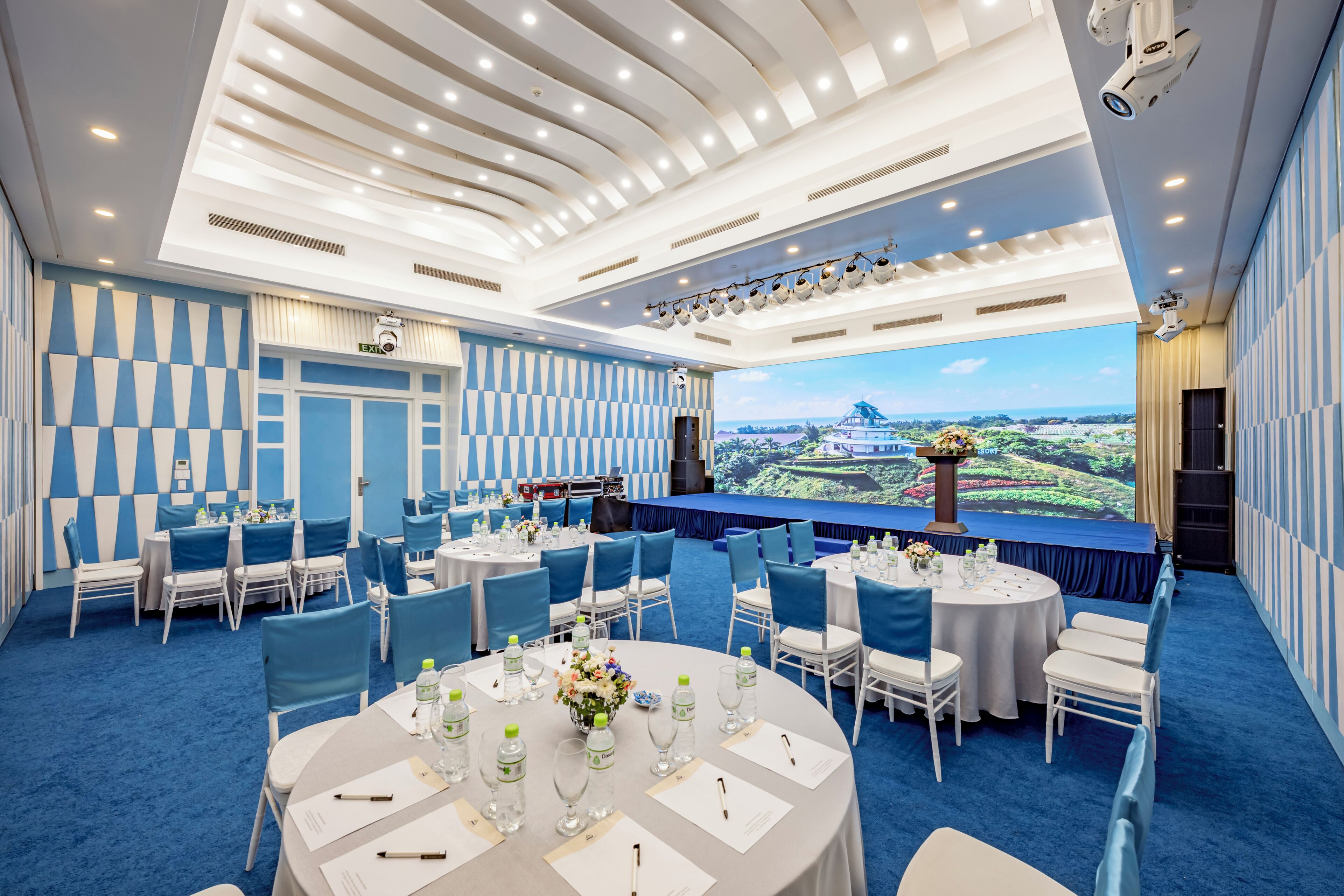 A conference room with round tables, chairs, and a stage with a scenic backdrop.