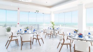A bright, spacious dining area with multiple round tables and white chairs, large windows offering a view of the ocean, and a ceiling with a decorative light fixture.