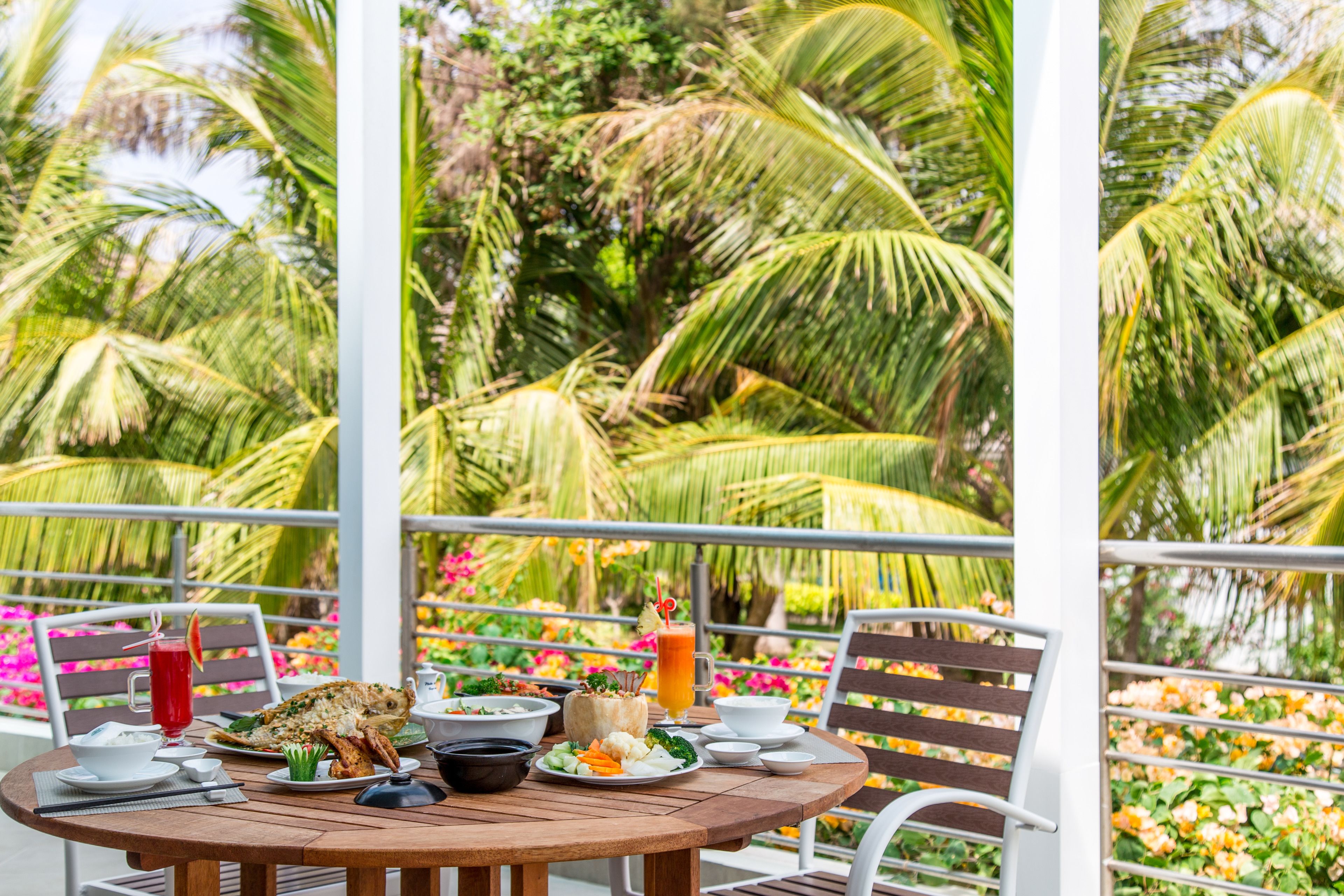 Una mesa redonda de madera con una comida dispuesta, que incluye pescado, verduras y bebidas, rodeada de sillas y una barandilla con vistas a palmeras y flores.