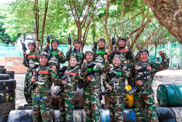 A group of people in military-style camouflage uniforms holding paintball guns, standing in an outdoor area with trees and barrels in the background.