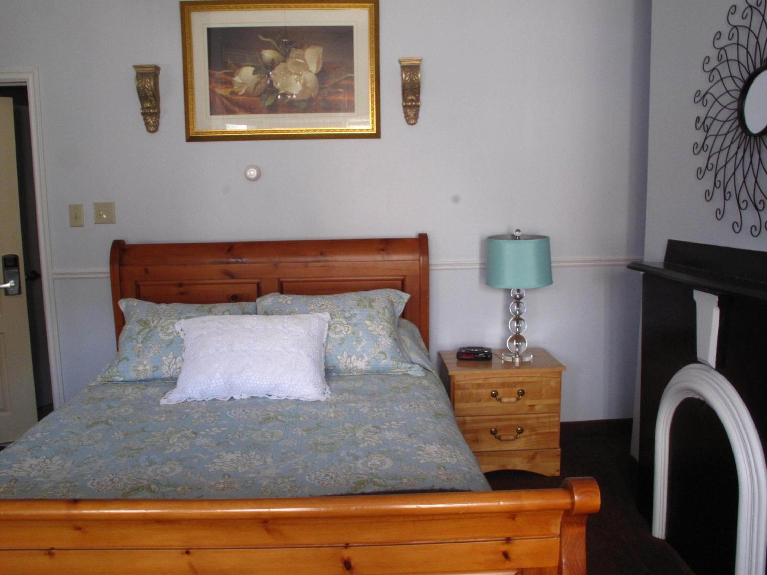 A neatly made bed with floral bedding, a wooden headboard, a bedside table with a lamp, and a wall-mounted picture.