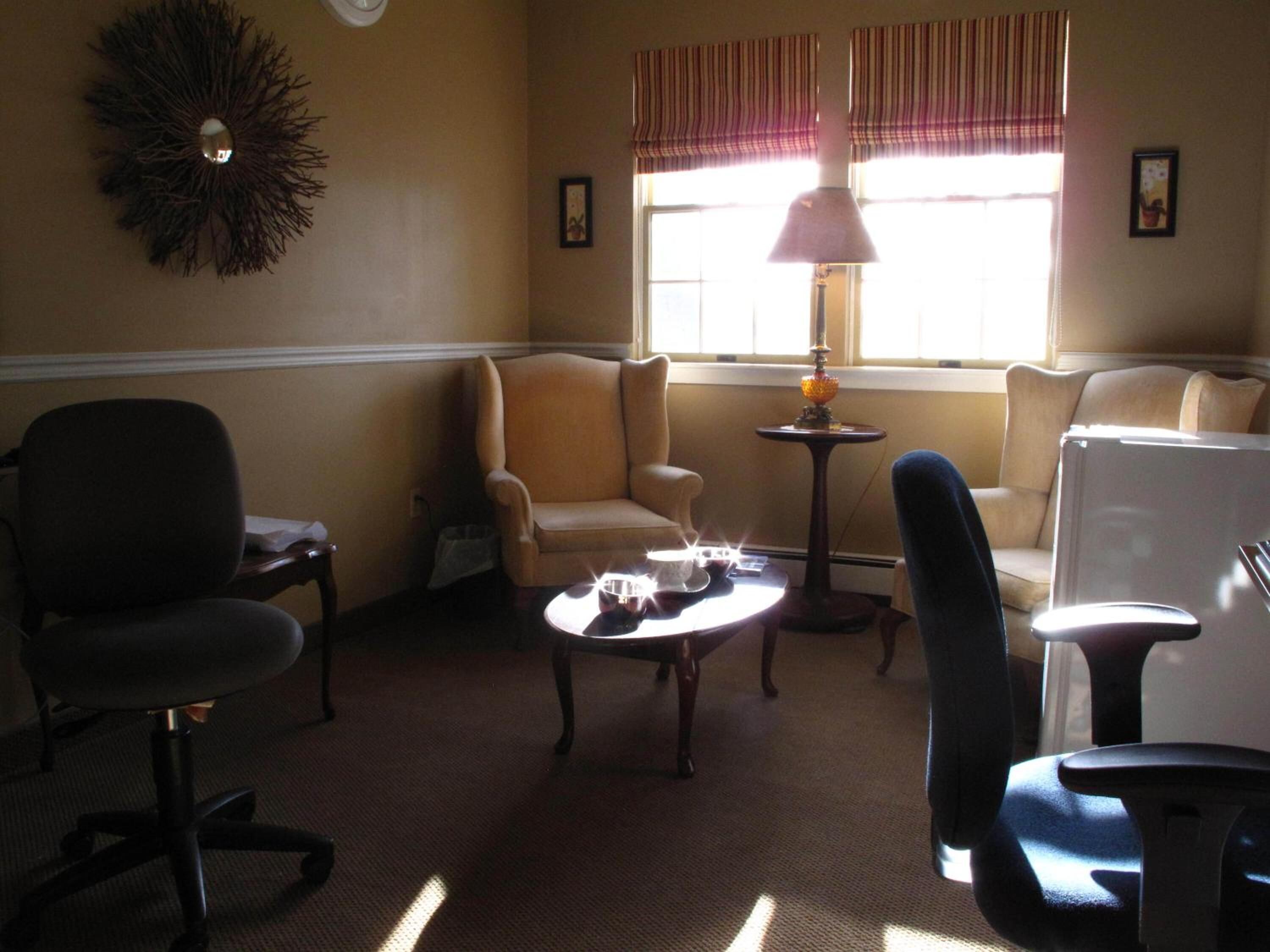 A room with a beige armchair, a black office chair, a small round table, and a lamp.