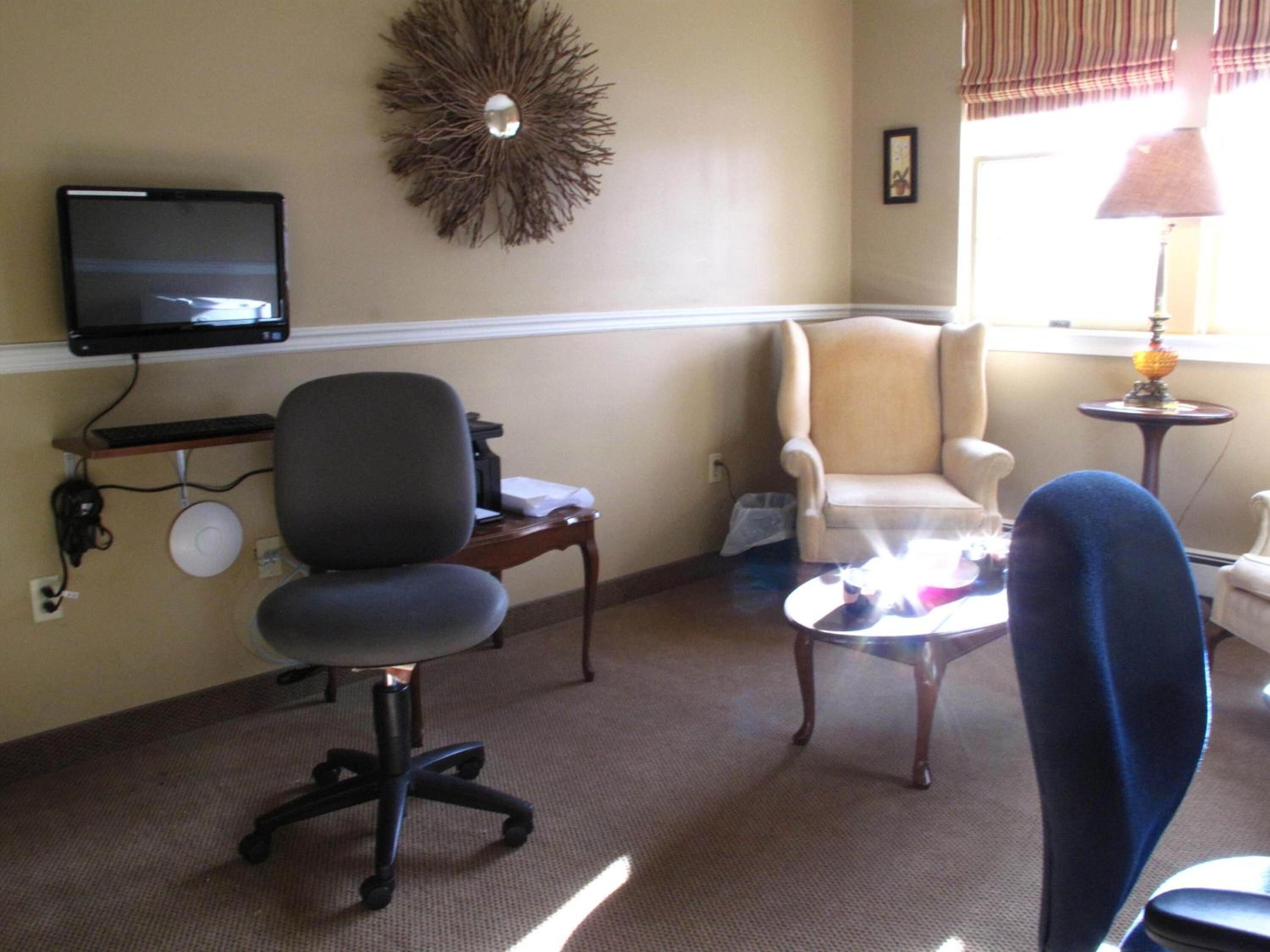 A room with a desk, office chair, armchair, and a small table with a lamp.