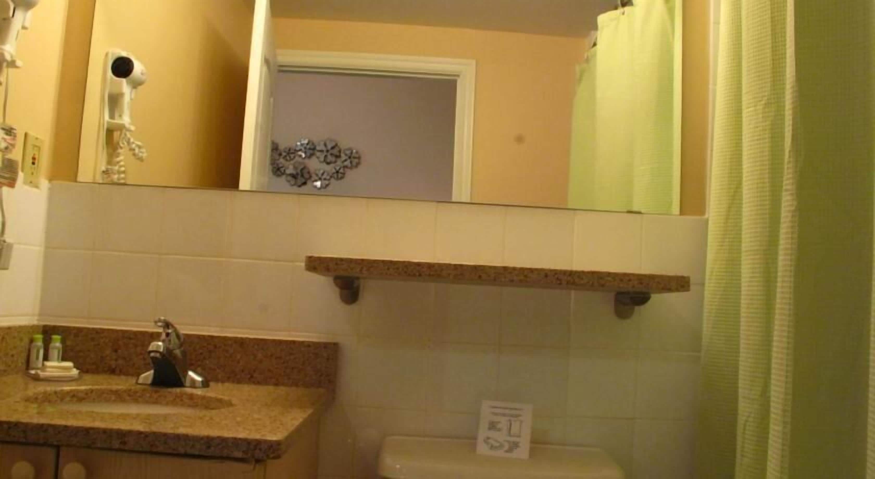 A bathroom with a sink, mirror, and toilet. There is a shelf above the toilet and a shower curtain.