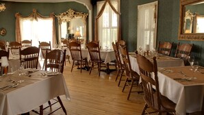 A dining area with multiple tables and chairs, wooden flooring, and large windows with curtains.