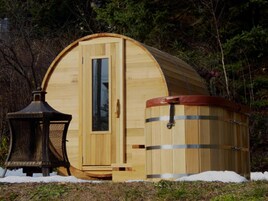 Una estructura de sauna de madera con una puerta grande y otra más pequeña, rodeada de nieve y árboles.