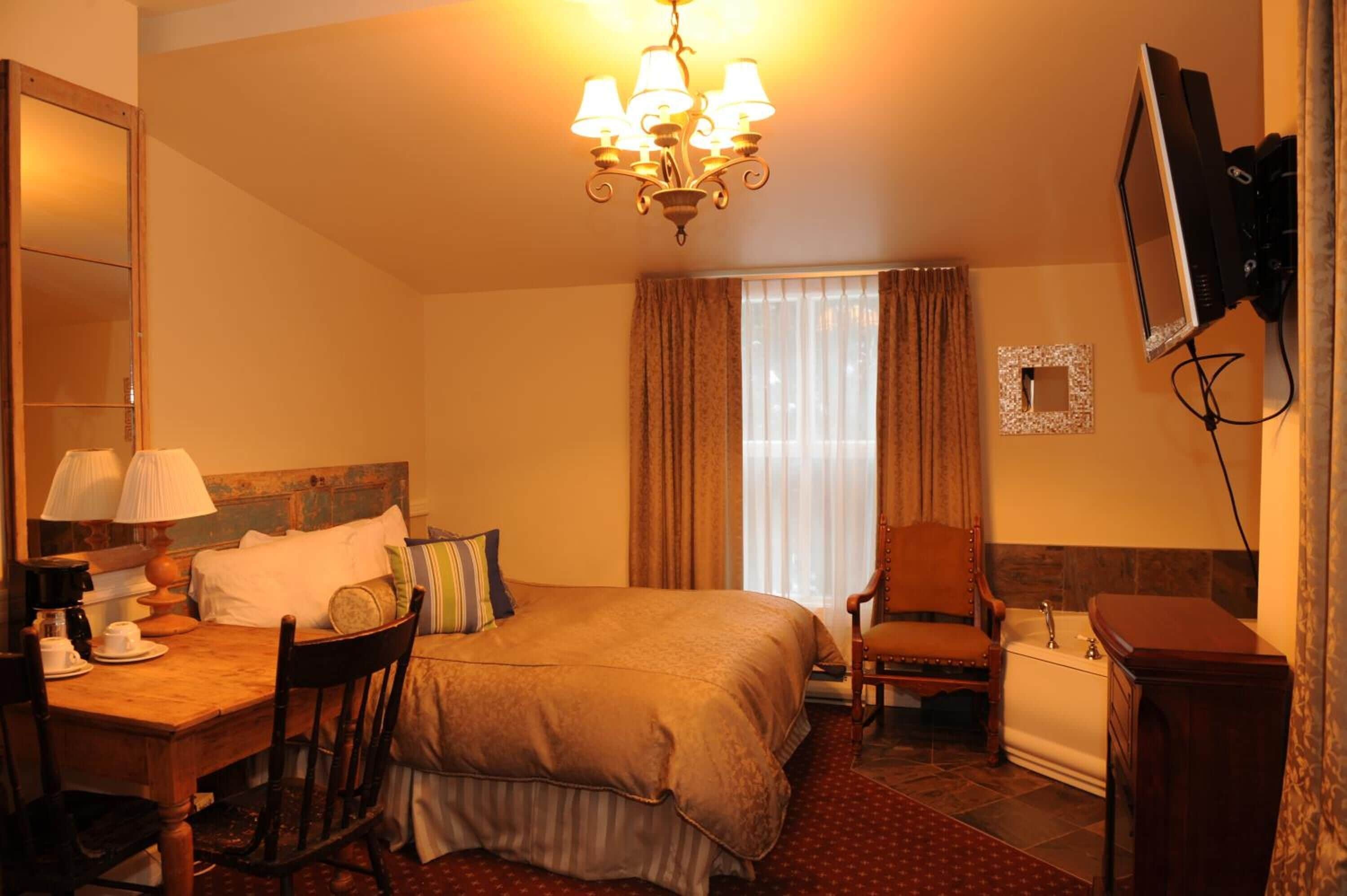 Habitación de hotel con cama, escritorio con lámpara, silla, televisor montado en la pared y ventana con cortinas.