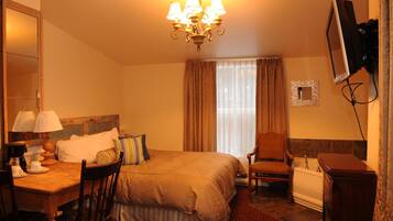 Habitación de hotel con cama, escritorio con lámpara, silla, televisor montado en la pared y ventana con cortinas.