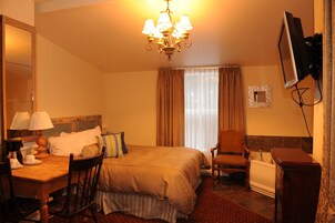 Habitación de hotel con cama, escritorio con lámpara, silla, televisor montado en la pared y ventana con cortinas.