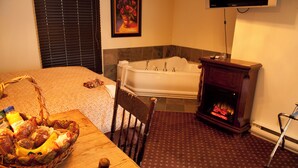 A hotel room with a bed, a wooden table with a fruit basket, a fireplace, and a large window with blinds.
